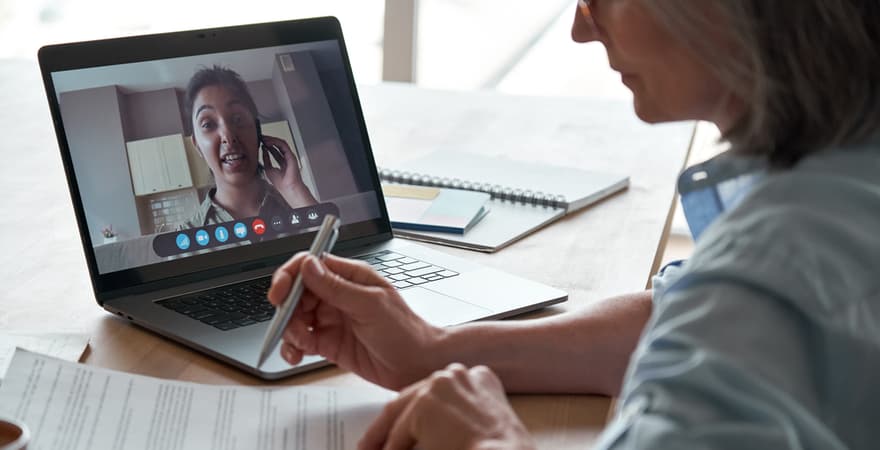 A counsellor sits at her desk making notes, while a client is talking on her laptop via video.