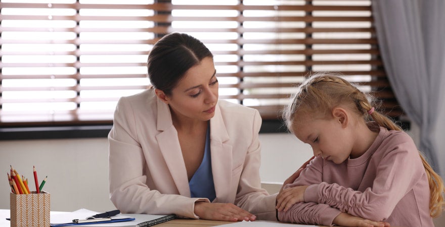 A counsellor sits in a classroom talking to a child who looks sad.