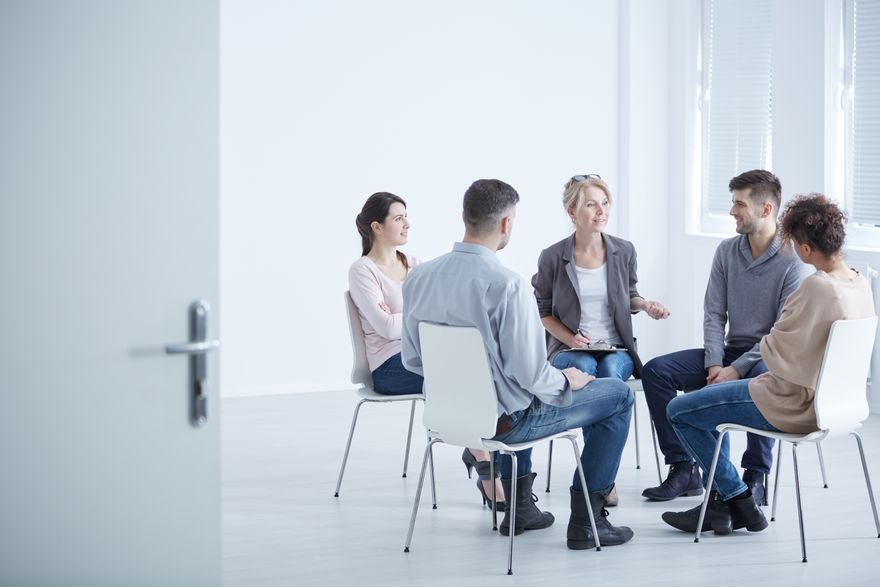 A group of people sit on chairs in a circle. A counsellor is talking and holding a clipboard, the others are listening. 