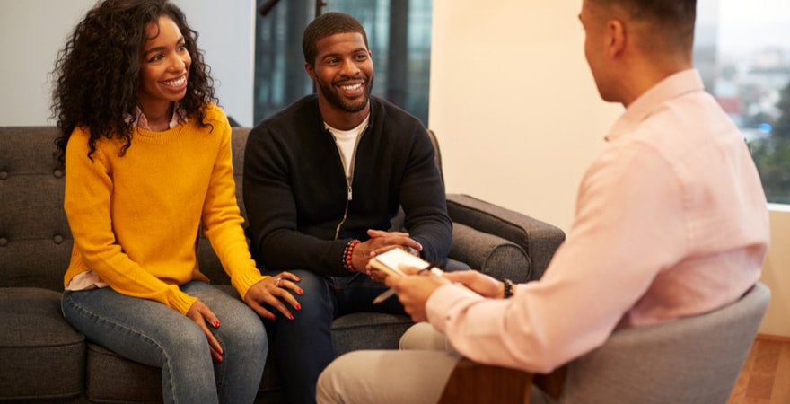 A couple are sitting on a couch, talking to a counsellor and smiling.