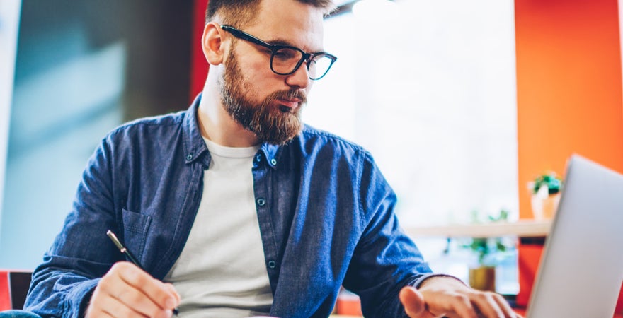 An ECU Online student sits at a table, writing in a notebook and checking his laptop.