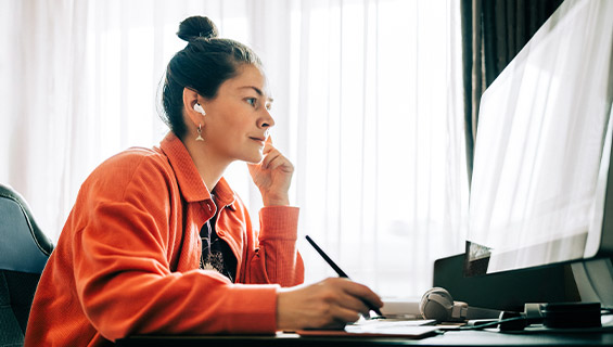 A woman sitting at a desk, working on a computer with a pen in hand, focused on her task.