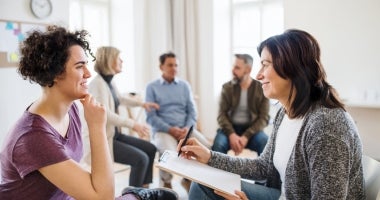 A woman sits in her chair, tlking to a counsellor. They are both smiling and there are people sitting and talking in the background.